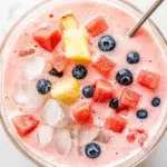 A glass bowl of Korean hwachae fruit punch filled with pink milky soda, watermelon cubes, blueberries, pineapple chunks, and ice.