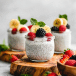 Creamy chia seed yogurt in small glass jars topped with banana slices, raspberries, blackberries, and fresh mint on rustic wooden boards.