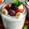 Creamy chia seed yogurt in glass jars topped with blackberries, strawberries, and banana slices on a wooden board.