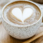 Hojicha Latte: The Rich & Toasty Japanese Drink Taking Over Cafes 5 Hojicha latte with heart latte art in a speckled ceramic cup and saucer with a wooden spoon on a checkered cloth.