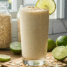 Tall glass of Oatzempic drink topped with oats and lime slice, with limes, oat flakes, and bottles in a cozy kitchen setting.