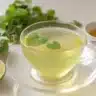 A clear glass teacup filled with lemon balm tea for weight loss recipe, surrounded by fresh lime slices, mint leaves, and honey on a white countertop.