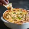 Buffalo Chicken Dip in a white baking dish with melted cheese and a chip being dipped into the gooey, cheesy mixture.