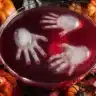 Halloween punch bowl with red punch and floating ice hands, surrounded by pumpkins and spooky fall decorations.