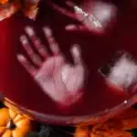 Halloween Punch with spooky red liquid and floating frozen hand-shaped ice cubes, surrounded by pumpkins and autumn leaves.