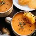 Rustic ceramic mugs filled with creamy butternut squash tomato soup, garnished with thyme and served with crispy parmesan crisps.