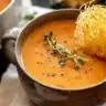 A cozy mug of butternut squash tomato soup garnished with thyme and a parmesan crisp, served on a dark rustic table with a linen napkin.