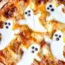 Homemade Halloween ghost pizza topped with melted mozzarella shaped like ghosts and black olive eyes on a wooden table.