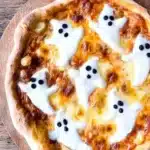 Homemade Halloween ghost pizza topped with melted mozzarella ghosts and black olive eyes on a wooden board.