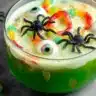 Green Halloween punch in a glass bowl decorated with gummy worms, floating eyeball candies, and plastic spiders.