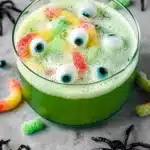 Green Halloween punch in a glass bowl decorated with gummy worms, candy eyeballs, and plastic spiders.