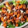 Colorful roasted sweet potato goat cheese salad with leafy greens, pecans, mandarin slices, and dried cranberries on a white serving platter.