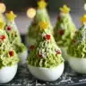 Deviled eggs topped with piped green filling shaped like Christmas trees, decorated with red peppers and cheese, each with a small star on top.