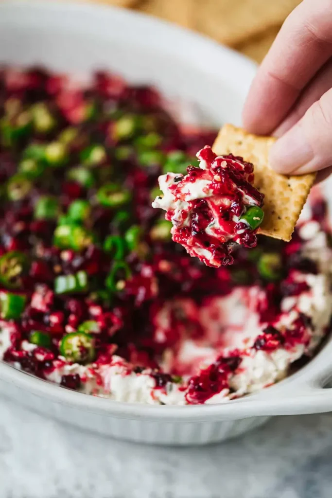Cranberry jalapeño cream cheese dip scooped with a cracker showing layers of cream cheese, cranberry relish, and jalapeños.