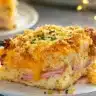 A slice of Christmas breakfast casserole layered with ham, cheese, fluffy bread, and a crispy golden topping served on a white plate.
