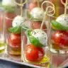 Mini Caprese bites in clear cups with cherry tomatoes, basil, and mozzarella skewered on knot picks and drizzled with olive oil.