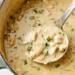Creamy white chicken chili soup in a Dutch oven with a ladle lifting tender chicken pieces, beans, and fresh herbs.