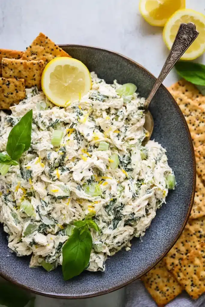 Bowl of creamy lemon basil chicken salad topped with fresh basil, surrounded by crackers and lemon slices.