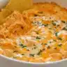 Buffalo chicken dip in a white dish topped with melted cheese and herbs with tortilla chips dipped inside.